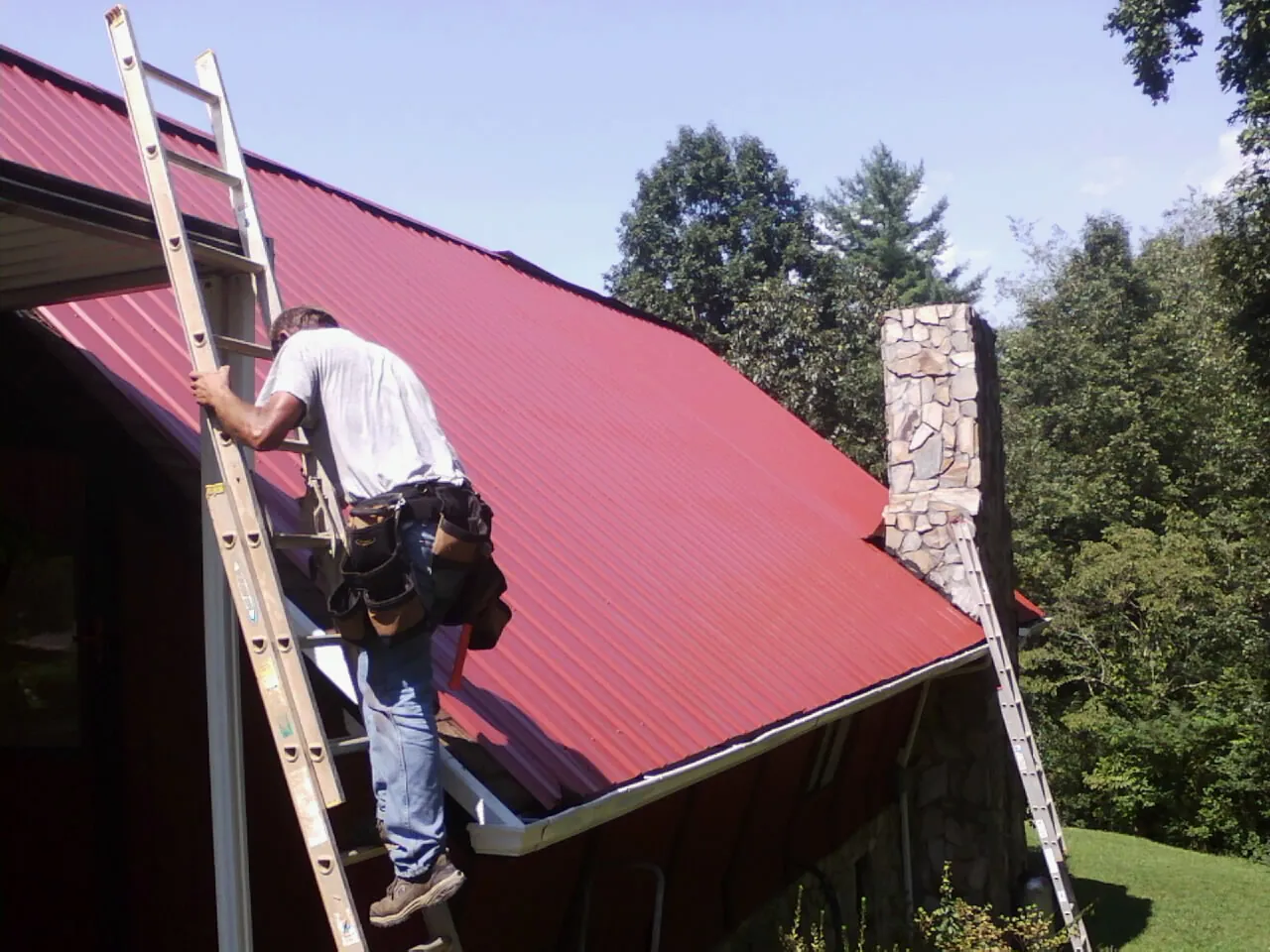 Roofer on a ladder beside a red metal roof and stone chimney — Soffit & Fascia Repair by Roofing Brothers in Conyngham